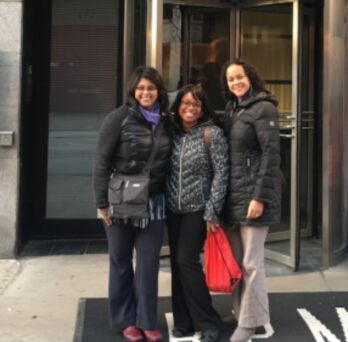three women wearing winter coats and standing outside a building entrance
                  